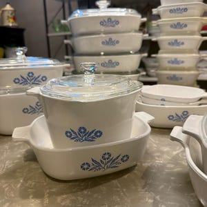 May include: A collection of white glass bakeware featuring a blue floral pattern. The set includes various rectangular and square casserole dishes with integrated handles and clear glass lids, showcasing a consistent design across all pieces.