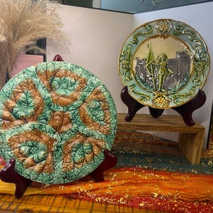May include: Two decorative ceramic plates on wooden stands. One plate features a leaf design in green and brown. The other plate has a castle scene with a figure holding a flag, in shades of green, blue, and gray.