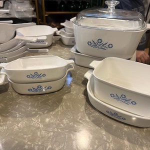 May include: A collection of white ceramic bakeware pieces featuring a blue floral pattern. The set includes various rectangular and square baking dishes with integrated handles, some stacked, and one large rectangular dish with a clear glass lid.