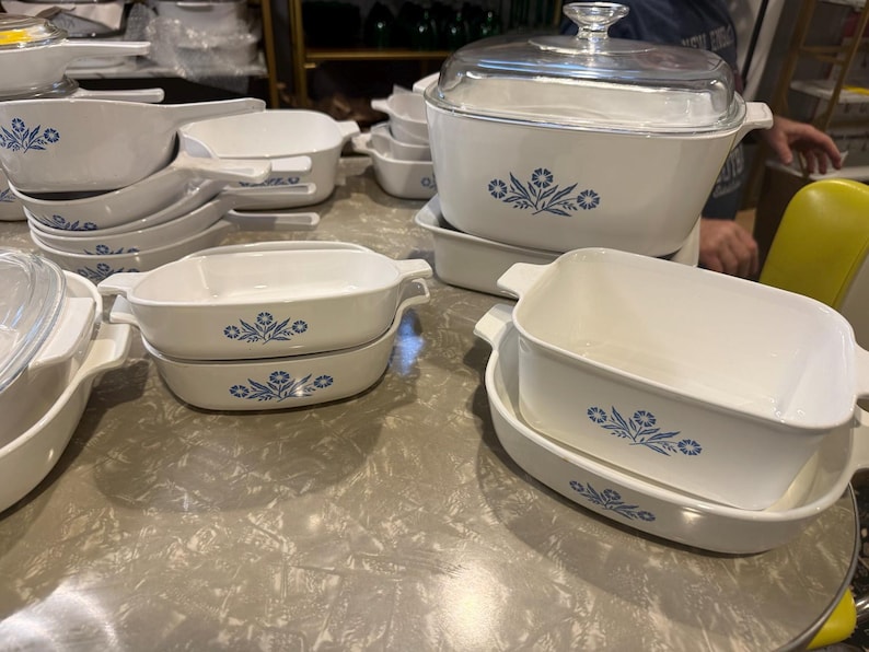 May include: A collection of white ceramic bakeware pieces featuring a blue floral pattern. The set includes various rectangular and square baking dishes with integrated handles, some stacked, and one large rectangular dish with a clear glass lid.