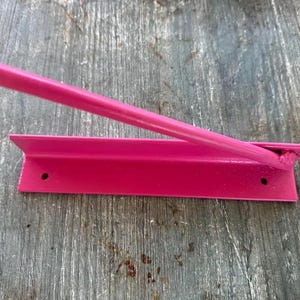 May include: A bright pink metal tool featuring a long, cylindrical handle attached to a flat, L-shaped base. The base includes two circular mounting holes on the horizontal flange, and the entire object is finished in a solid, vibrant pink paint.