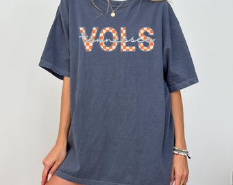 May include: A slate blue short-sleeve t-shirt featuring the word VOLS in an orange and white checkered pattern, with the word tennessee written in white cursive script across the center. The shirt has a crew neckline and a relaxed fit.