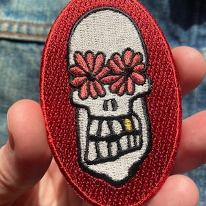 Può includere: Toppa ricamata ovale con un teschio. Il teschio ha occhi a forma di fiore e un dente d'oro, su sfondo rosso. La toppa è probabilmente per abbigliamento o accessori.