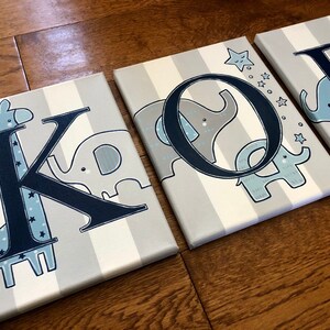 May include: Canvas art pieces spelling out the name "KOB". Each canvas features a letter in navy blue with a corresponding animal illustration in light blue and gray. The background has vertical gray and white stripes.