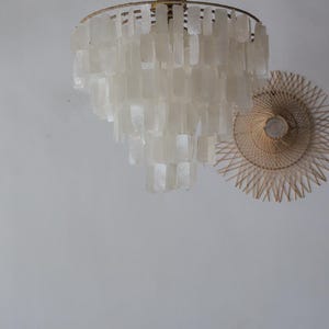 May include: A tiered chandelier with rectangular, off-white shell-like pieces hangs from a gold-colored metal frame. A round, woven wall decoration with a light bulb in the center is to the right of the chandelier. The background is a plain, light-colored wall.