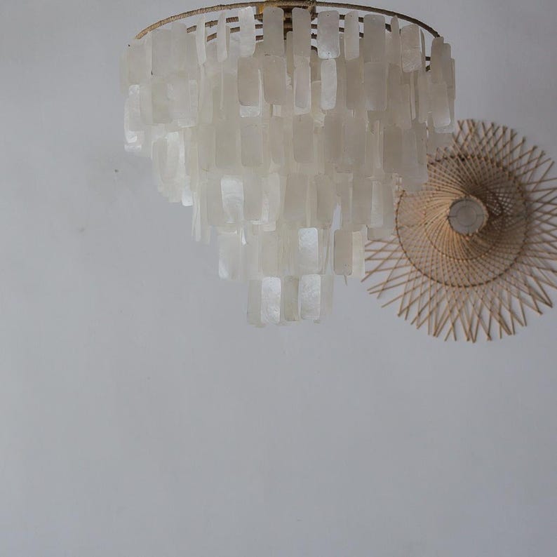 May include: A tiered chandelier with rectangular, off-white shell-like pieces hangs from a gold-colored metal frame. A round, woven wall decoration with a light bulb in the center is to the right of the chandelier. The background is a plain, light-colored wall.