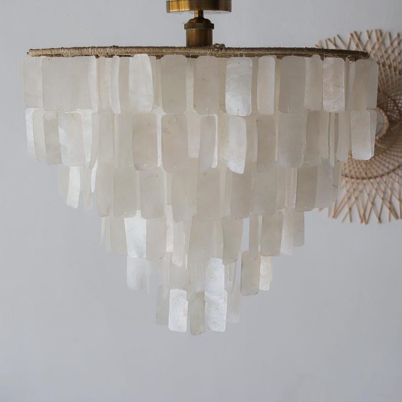 May include: A tiered chandelier with a brass fixture and a cascade of rectangular, off-white shell pieces. The light fixture is suspended from the ceiling. A woven wall decor is visible in the background.