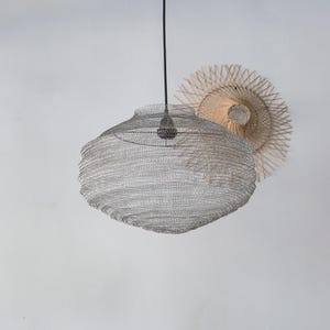 Puede incluir: Una lámpara colgante de malla de alambre plateada con una bombilla. La luz tiene una forma redondeada y texturizada. En el fondo hay una decoración de pared en forma de sol tejida.
