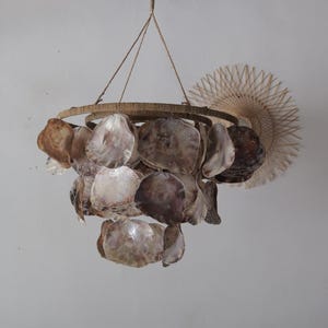 Könnte beinhalten: Dekorativer Kronleuchter aus natürlichen Capiz-Muscheln in Braun-, Lila- und Weißtönen. Die Muscheln sind an einem kreisförmigen Rahmen befestigt und an einem Seil aufgehängt. Im Hintergrund ist ein gewebter Fächer zu sehen.