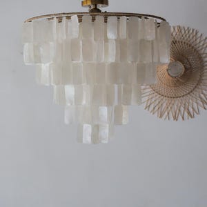 May include: A tiered chandelier with rectangular, off-white shell-like pieces. The fixture has a gold-colored metal frame and hangs from a matching ceiling mount. A woven, circular wall decoration is visible in the background.