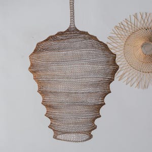 以下が含まれることがあります： ユニークな層状のデザインの、ブロンズ色の織り金属ペンダントライト。 ランプは、質感のある表面と吊り下げ用の細い上部を備えた球根状の形状をしています。 背景には、2つ目の円形の織りライトフィクスチャが見えます。