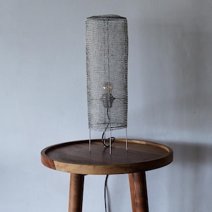 May include: A cylindrical table lamp with a metal mesh shade. The lamp has a visible lightbulb and stands on thin metal legs. It is placed on a round wooden side table.