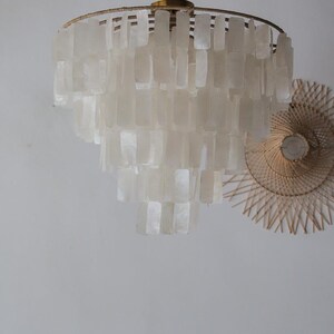 May include: A tiered chandelier with a gold-colored frame and numerous rectangular, off-white shell-like pieces. The light fixture is suspended from the ceiling. A woven, circular wall decoration is visible in the background.