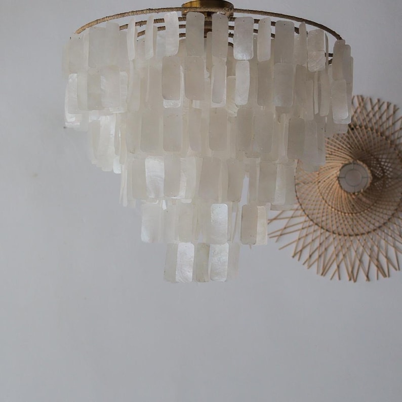 May include: A tiered chandelier with a gold-colored frame and numerous rectangular, off-white shell-like pieces. The light fixture is suspended from the ceiling. A woven, circular wall decoration is visible in the background.