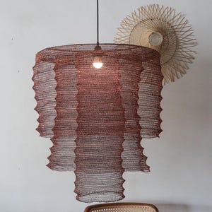 Pode incluir: Uma luminária pendente de vários níveis feita de malha de arame na cor cobre escuro. A estrutura apresenta três camadas cilíndricas distintas e sobrepostas de larguras variáveis, suspensas por um fio preto com uma lâmpada central acesa.