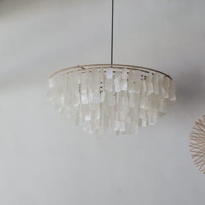 May include: A round, hanging chandelier with a natural woven frame and cascading, rectangular, white shell accents. The light fixture is suspended from a black cord against a neutral wall. The design evokes a coastal or bohemian aesthetic.