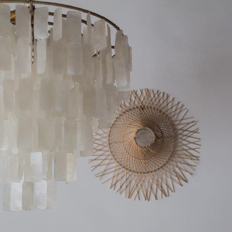 May include: A close-up of two light fixtures. One is a chandelier with rectangular, off-white shell-like pieces. The other is a round, woven, natural-colored fixture with a central light source. Both are against a white background.