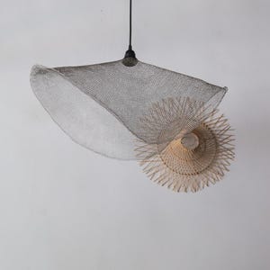 May include: A modern pendant light fixture featuring a large, silver wire mesh shade above a smaller, natural woven shade. The light hangs from a black cord and fixture against a white background.