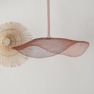 May include: A copper-colored, wave-shaped pendant light fixture made of woven metal mesh. The light hangs from a chain and is suspended in front of a white wall. A woven, circular wall decoration is to the left of the light.