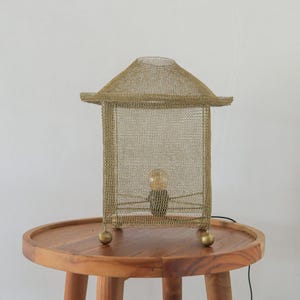 Könnte beinhalten: Eine goldfarbene Metallgitter-Laternenlampe mit einer kleinen Glühbirne im Inneren. Die Lampe hat einen rechteckigen Körper und ein schräges Dach. Sie steht auf einem runden Holztisch mit drei Beinen. Der Lampenfuß hat vier kleine, runde Füße.