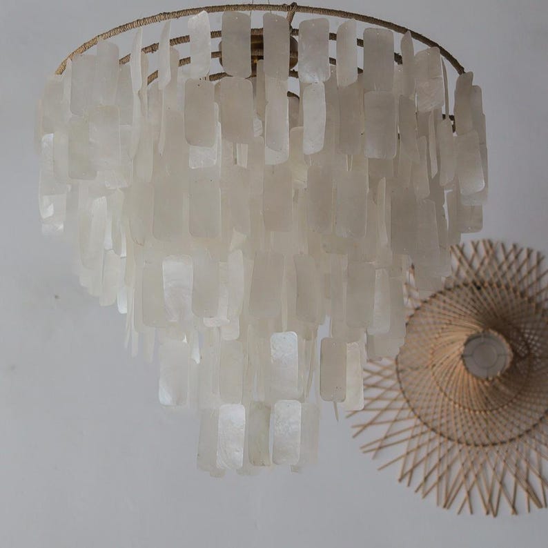 May include: A tiered, off-white chandelier with rectangular shell-like pieces hanging from a circular frame. The chandelier is suspended from the ceiling. A woven, circular wall decor is visible in the background.