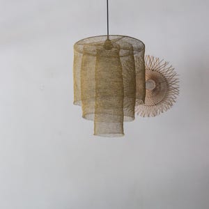 May include: A gold-coloured, tiered pendant light fixture with a mesh design hangs from a black cord. The light has a cylindrical shape with three descending tiers. A woven, circular wall decoration is visible in the background.