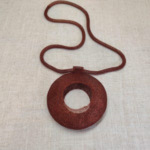 May include: A reddish-brown, woven metal necklace with a circular pendant and a matching cord. The pendant has a hollow center and a textured surface, creating a unique, eye-catching accessory. The necklace is displayed on a neutral background.