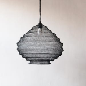 Puede incluir: Una lámpara colgante de malla de alambre negro con una forma redondeada y orgánica. La lámpara está colgada de un cable negro.