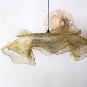May include: A gold mesh pendant light with a flowing, cloud-like design. The light hangs from a black cord against a white background. A woven, circular element is visible above the light, adding a natural touch.
