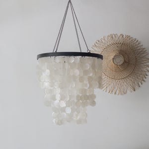 Op de afbeelding: Een witte schelp kroonluchter met een zwart metalen frame en ketting. De kroonluchter hangt aan het plafond en heeft meerdere lagen schelpen.