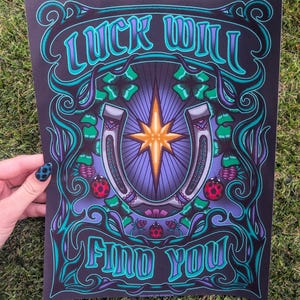 May include: A rectangular art print on a black background featuring the text LUCK WILL FIND YOU in stylized teal and purple lettering. The design includes a central silver horseshoe, a glowing orange star, green four-leaf clovers, and ladybugs.