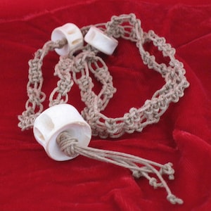 May include: A handmade macrame bracelet with three white, cylindrical beads. The bracelet is made of beige-colored hemp cord and is displayed on a red velvet surface. The beads have a carved design.