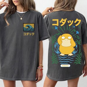 May include: A charcoal gray t-shirt featuring a small graphic on the front chest of a blue wave with yellow text. The back displays a large yellow duck-like character, green palm leaves, blue stars, and the Japanese text for Psyduck.