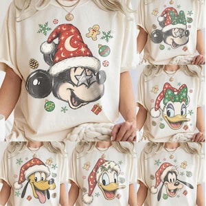 Peut inclure: Un t-shirt à manches courtes de couleur crème arborant un motif de Mickey Mouse portant un bonnet de Père Noël rouge à motifs de lunes et d'étoiles, des lunettes en forme d'étoile, entouré de bonshommes en pain d'épice, d'ornements verts et de flocons.