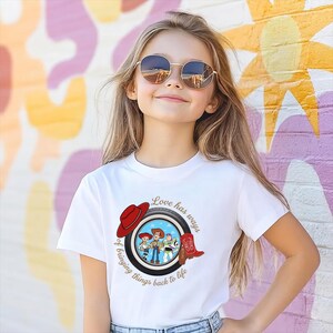 May include: A white short-sleeved t-shirt featuring a graphic of a tire framing characters Jessie, Woody, and Buzz Lightyear. The design includes a red cowboy hat, a red cowboy boot, and the text Love has ways of bringing things back to life.