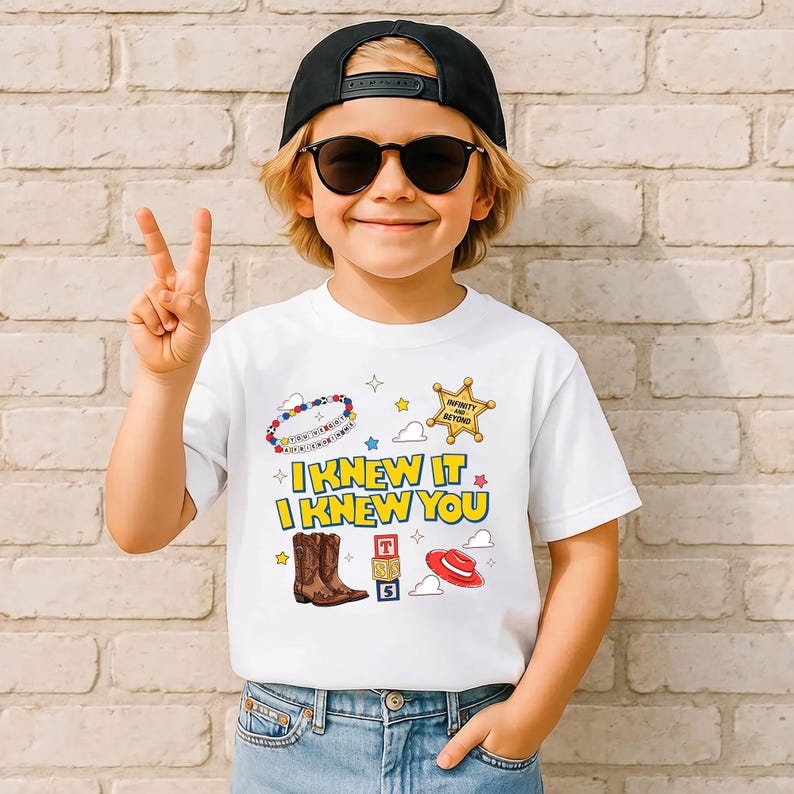 May include: A white short-sleeve t-shirt featuring a graphic print with the text "I KNEW IT I KNEW YOU" in yellow and blue. The design includes illustrations of brown cowboy boots, a red hat, a sheriff badge, alphabet blocks, and a beaded bracelet.