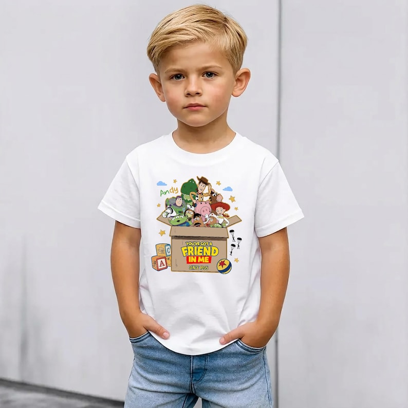 May include: A white short-sleeved t-shirt featuring a graphic of Toy Story characters emerging from a cardboard box. The box is labeled with the text 'YOU'VE GOT A FRIEND IN ME SINCE 1995' and the name 'Andy' is written in green above the box.