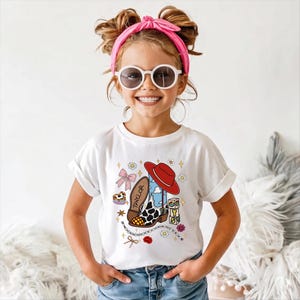 May include: A white short-sleeved t-shirt featuring a graphic print of a cowboy boot with cow print and blue sky patterns, a red cowboy hat, a pink bow, small flowers, stars, and blocks spelling TAYLOR. The design includes a beaded chain element.