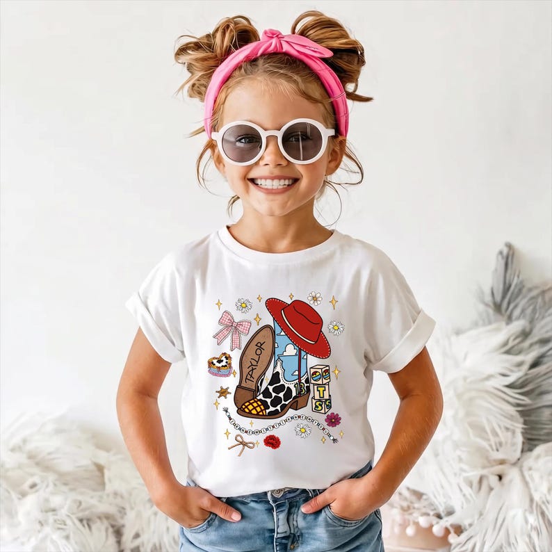 May include: A white short-sleeved t-shirt featuring a graphic print of a cowboy boot with cow print and blue sky patterns, a red cowboy hat, a pink bow, small flowers, stars, and blocks spelling TAYLOR. The design includes a beaded chain element.
