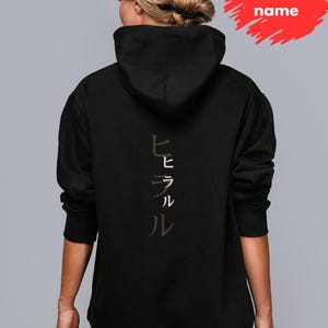 May include: Black hooded sweatshirt with Japanese characters printed vertically down the back. The text is in a muted, earthy tone. The sweatshirt has a drawstring hood and long sleeves. The image also includes a red banner with the text "Your Japanese name".
