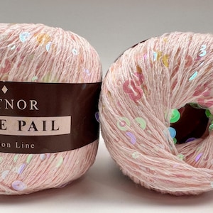 以下が含まれることがあります： ピンク色の糸玉が2つ。キラキラ光るスパンコール付き。「Kutnor Shine Pail Fashion Line」のラベル。糸は薄いピンク色で、カラフルなスパンコールが散りばめられています。1つの玉は部分的にほどかれています。