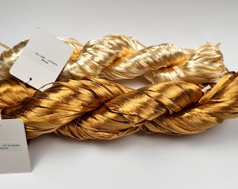 May include: Two skeins of metallic gold yarn, one in a darker shade and the other a lighter, blonde tone. The yarn is twisted and braided, with white tags attached. The tags contain text.