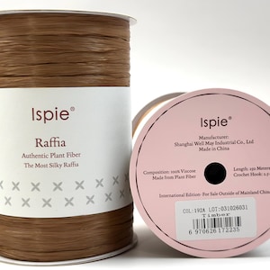 May include: A spool of brown Ispie raffia made from 100% viscose plant fiber. The label reads Ispie Raffia Authentic Plant Fiber, and the side label indicates a length of 250 meters, a crochet hook size of 2.5-4mm, and the color Timber.