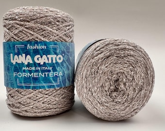 Può includere: Due gomitoli di filato Lana Gatto. Un gomitolo è in posizione verticale, l'altro è sul lato. Il filato è di colore marrone chiaro con una lucentezza metallica. L'etichetta recita "Lana Gatto Made in Italy Formentera."