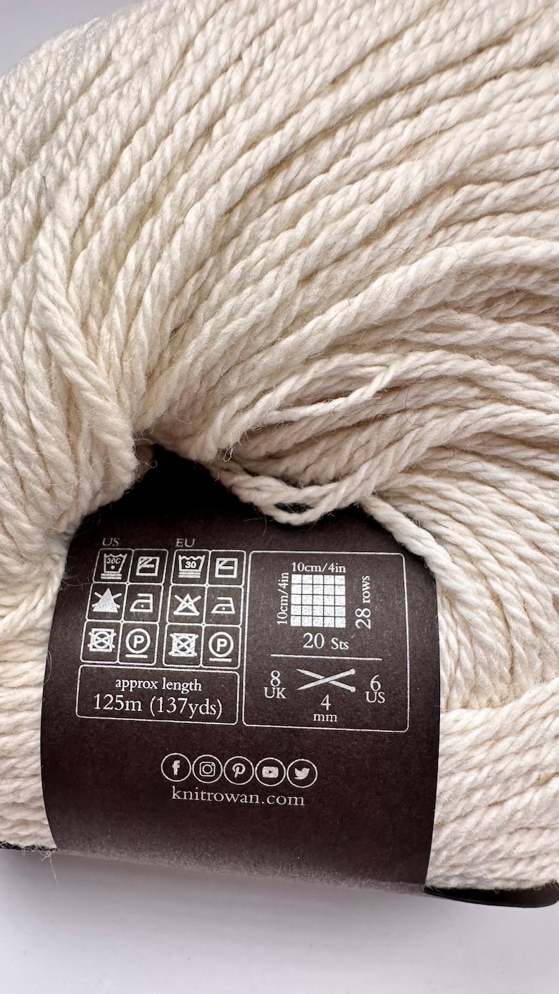 May include: A skein of off-white yarn with a black paper label. The label displays care instructions, a gauge of 20 stitches and 28 rows for a 4-inch square, a length of 137 yards, and a 4 mm needle size. The text knitrowan.com is printed at the bottom.