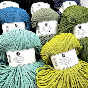 Puede incluir: Una colección de coloridas madejas de hilo de algodón reciclado en varios tonos, incluyendo azul, verde y amarillo. Cada madeja está etiquetada con el nombre del color y las especificaciones. El hilo está bien enrollado, mostrando su textura.