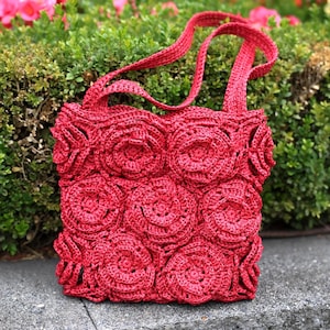May include: A red crocheted tote bag with a floral design. The bag features a series of large, circular flower patterns across its front and sides. It has two long straps for carrying over the shoulder. The bag is made of a textured, woven material.