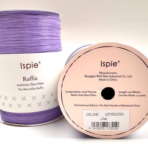 Puede incluir: Dos rollos de cinta de rafia de color lila. Un rollo está de pie, mostrando la marca "Ispie" y los detalles del producto. El otro rollo está tumbado, mostrando la etiqueta con información, incluyendo 150 metros de cinta.
