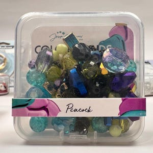 May include: A clear plastic container filled with a variety of colorful glass beads. The beads are in shades of blue, green, purple, and black. The container has a label that says "Peacock".