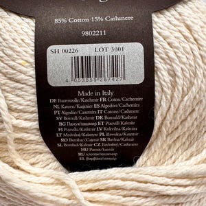 May include: A ball of off-white yarn with a dark brown paper label. The label states 50g, 85% Cotton 15% Cashmere, and Made in Italy. It includes a barcode, lot number 3001, and product code 9802211. The yarn has a thick, twisted texture.